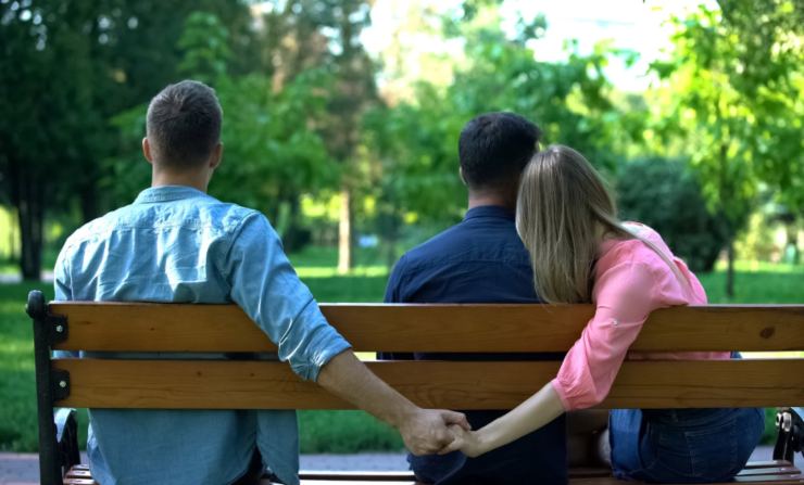 donna su una panchina abbracciata ad un uomo ma che da dietro dà la mano ad un altro uomo
